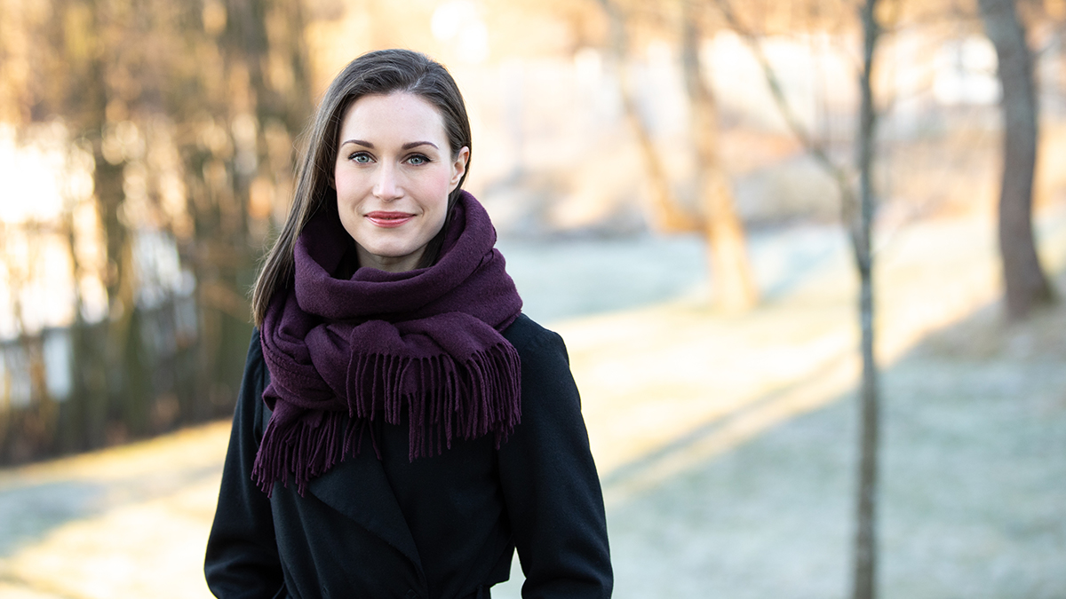 Pääministeri Sanna Marin seisoo ja hymyilee musta takki ja violetti kaulahuivi yllään.