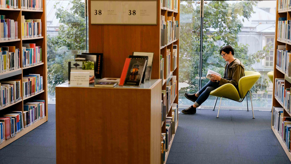 Nainen lukee kirjaa kirjastossa ikkunan äärellä