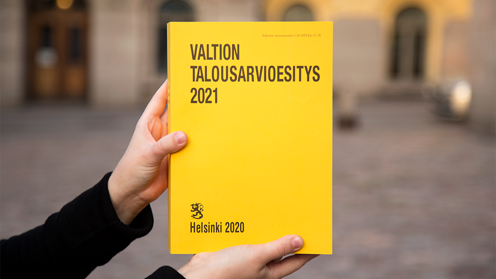 Kädet pitelevät vuoden 2021 budjettikirjaa Valtioneuvoston linnan edustalla.