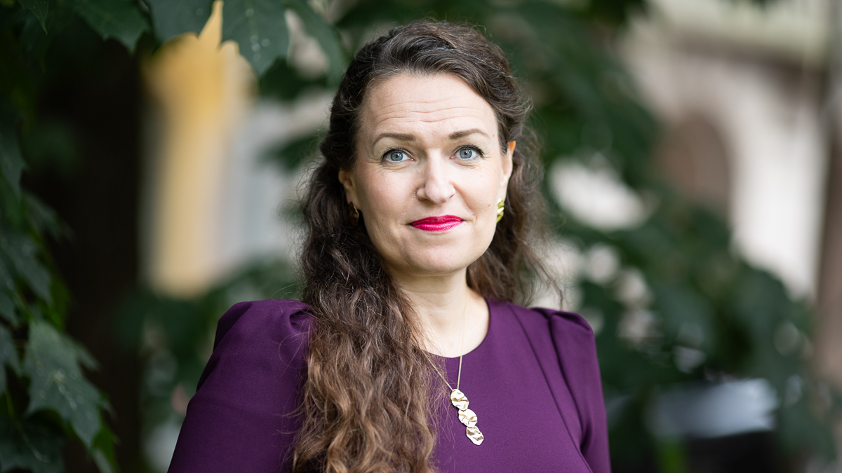 Ministeri Anna-Kaisa Ikonen kuvassa. Taustalla puu ja lehtiä.