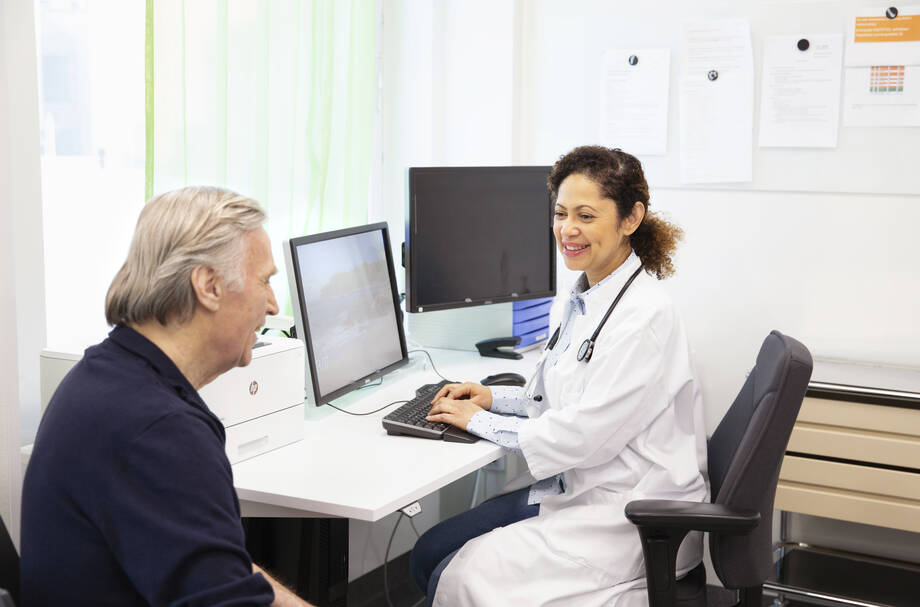 Patienten sitter på läkarens mottagning. Läkaren sitter vid datorn och matar patietens uppgifter in i patientdatasystemet.